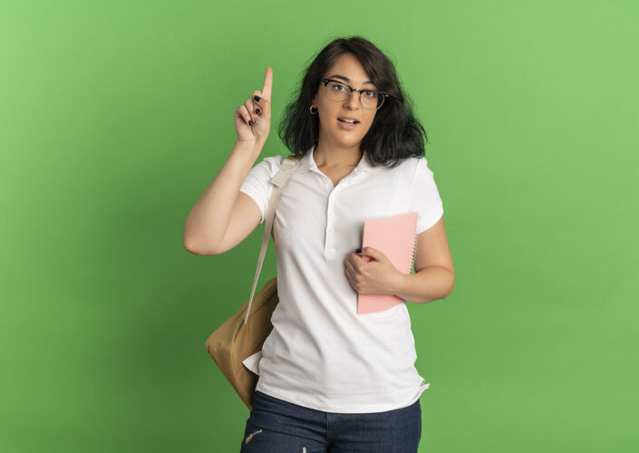 包年轻漂亮的白人女生戴着眼镜 背着书包 拿着笔记本指着绿色的笔记本 上面有复印空间笔记本惊喜绿色