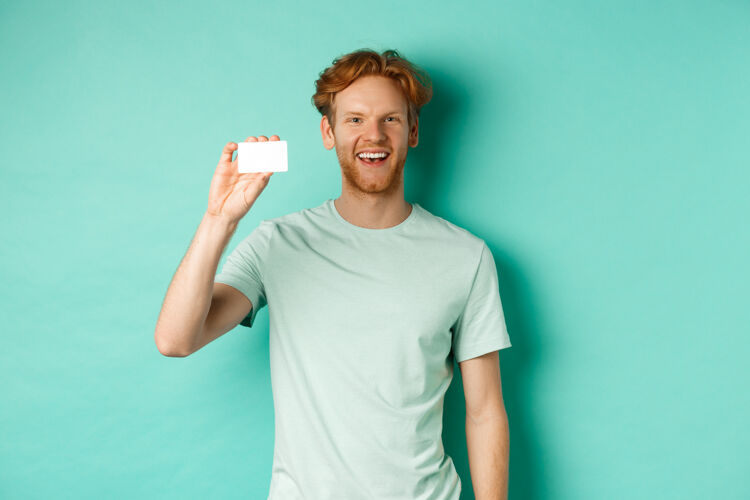成人购物理念穿着t恤的开朗的年轻人展示着塑料信用卡 微笑着站在薄荷背景下肖像姜家伙