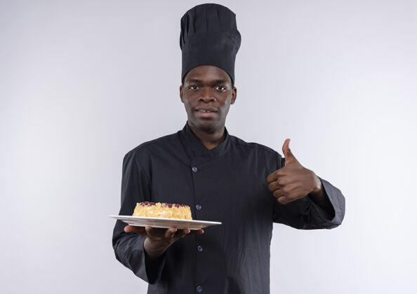 盘子身着厨师制服的年轻美籍黑人厨师手拿蛋糕放在盘子上 竖起大拇指放在白色餐具上大拇指蛋糕年轻