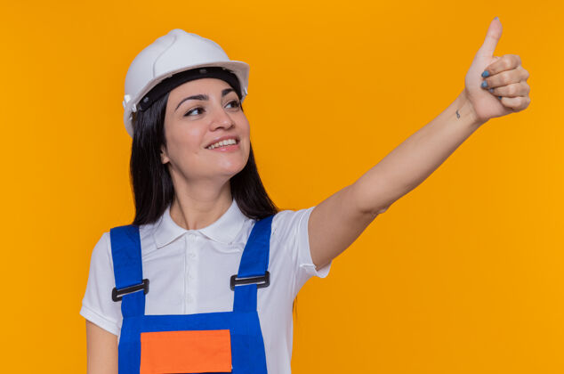 展示身穿施工制服 头戴安全帽的年轻建筑工人站在橙色的墙上 一边微笑一边自信地竖起大拇指安全制服年轻