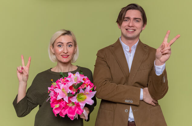 表演年轻漂亮的夫妇幸福的男人和女人拿着花束看着前面 微笑着兴高采烈地展示v形标志庆祝国际妇女节站在绿色的墙上快乐美丽花束