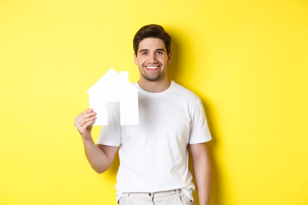 房地产房地产概念身穿白色t恤的年轻人拿着纸房子模型 微笑着 寻找公寓 站在黄色背景上表情模特自信
