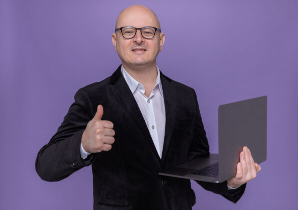 秃头身穿西装的中年秃头男子戴着眼镜拿着笔记本电脑望着前方微笑着兴高采烈地竖起大拇指站在紫色的墙上笔记本电脑拇指秀
