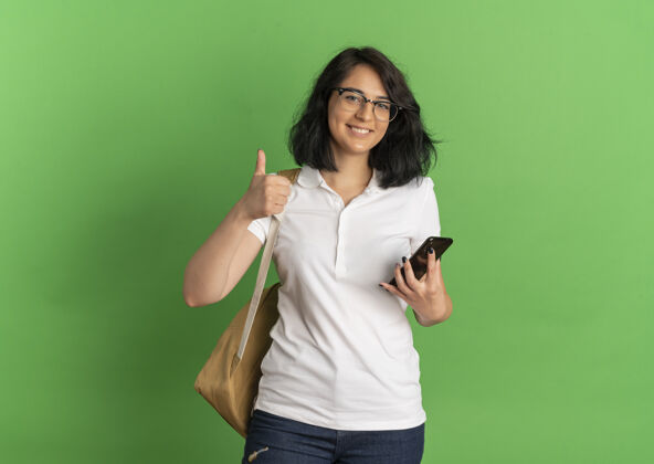 拇指年轻的高加索女孩 面带微笑 戴着眼镜 背着书包 竖起大拇指 拿着手机 放在绿色的空间里微笑包复制