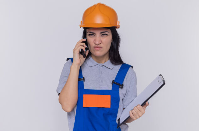 站着身穿建筑制服 戴着安全帽 拿着写字板的年轻建筑工人站在白色的墙上 一边用手机说话 一边显得很不高兴拿着年轻人女人