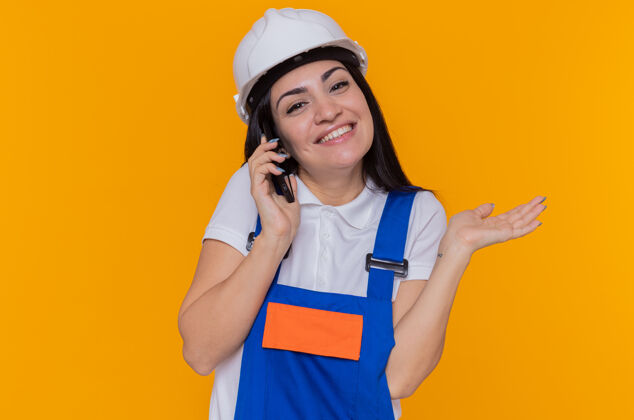 建设者身穿施工制服 头戴安全帽的年轻建筑工人站在橘色的墙上 一边微笑着举起手臂一边讲手机头盔手机电话