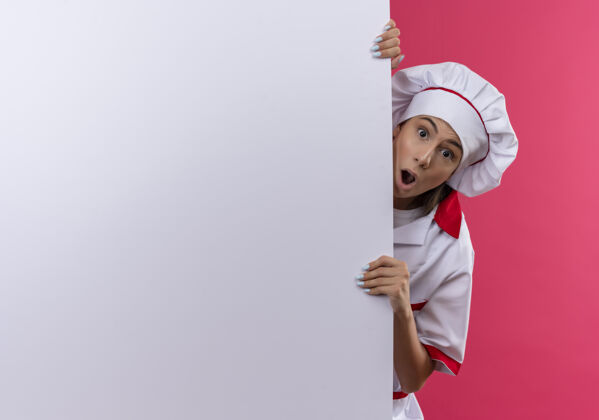 惊喜穿着厨师制服的年轻白人厨师女孩站在白色墙壁后面 粉色的墙上留有复印空间厨师复制烹饪