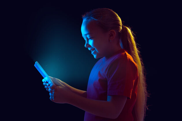 平板电脑微笑霓虹灯下隔离在黑暗墙上的白人女孩肖像使用平板电脑的美丽女性模特人类情感的概念 面部表情 销售 广告 现代科技 小玩意在线颜色工作