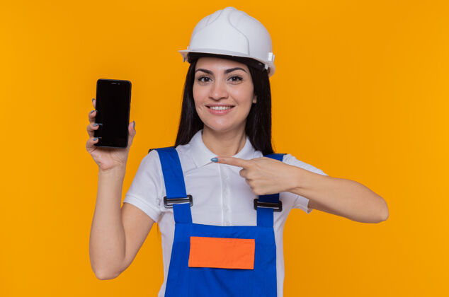 微笑身穿建筑制服 头戴安全帽的年轻建筑女工人站在橙色的墙上 用食指指着智能手机 开心地微笑着指点欢呼目录