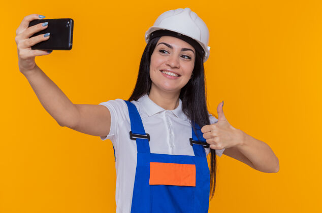 微笑身穿建筑制服 戴着安全帽的年轻建筑工人正在用智能手机自拍 自信地微笑着站在橙色的墙上竖起大拇指年轻头盔建筑