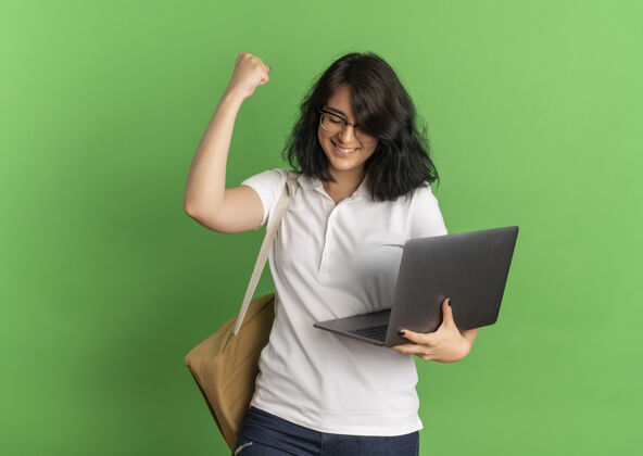 提高年轻笑容可掬的高加索女学生戴着眼镜 背着书包 手里拿着笔记本电脑 在绿色的空间里举起拳头年轻空间持有