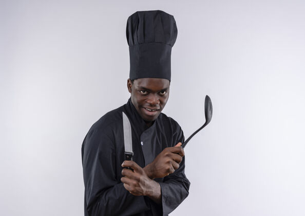 持有年轻快乐的非洲裔美国厨师穿着厨师制服 拿着刀和抹刀在白色的复制空间副本烹饪抹刀