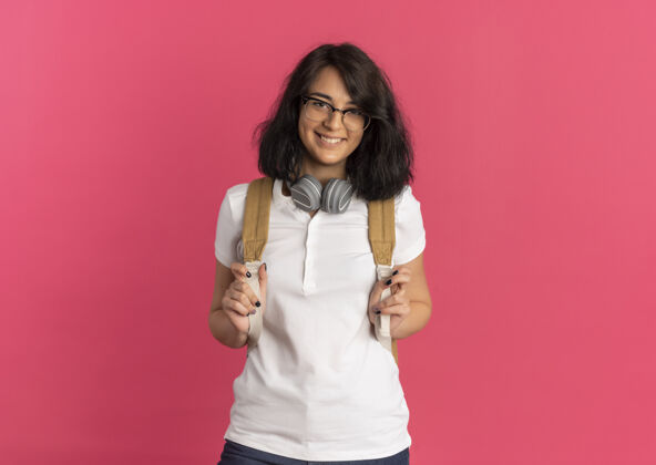 背部年轻的高加索女孩 戴着眼镜 戴着耳机 背着书包 面带微笑 看着粉红色的相机 留着复印空间脖子女学生穿