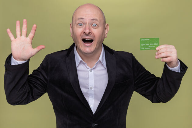 开穿着西装的中年秃头男人展示着信用卡的快乐和兴奋 展示着张开的五号手掌站在绿色的墙上手掌兴奋信用