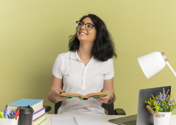 漂亮年轻笑容可掬的白种女孩戴着眼镜坐在书桌旁 手里拿着学习用具 抬起头来 手里拿着一本书放在绿色的小册子上空间向上眼镜