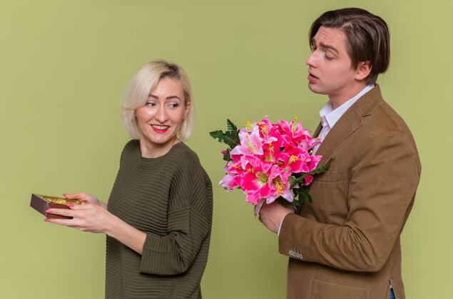 美丽年轻漂亮的情侣幸福的男人站在绿色的墙上 拿着一盒巧克力糖果 向微笑的女友送上一束鲜花 庆祝国际妇女节糖果花女人
