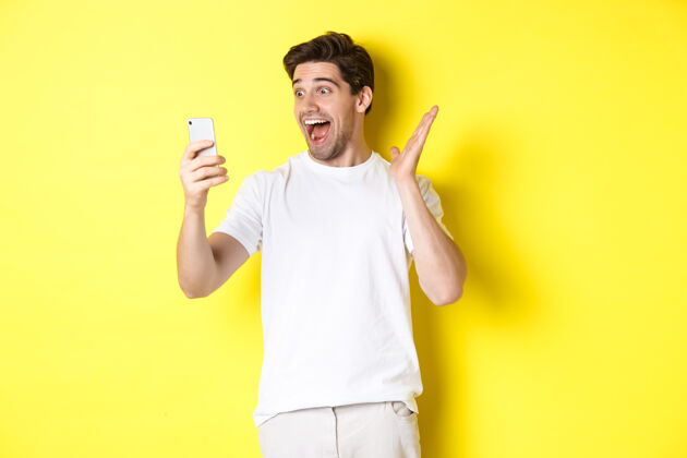 男人惊讶而快乐的男人看着手机屏幕 读着精彩的新闻 站在黄色的背景上快乐男人表情