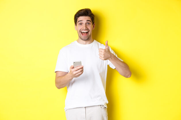工作室快乐的微笑的男人拿着智能手机 竖起大拇指表示赞同 在网上推荐一些东西 站在黄色的背景上男人成人男人