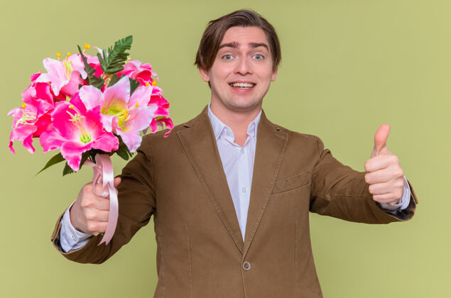 男人快乐的年轻人手持花束 微笑着 竖起大拇指 站在绿色的墙上 向国际妇女节表示祝贺祝贺欢呼花