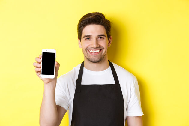 男士穿着黑色围裙的帅哥服务员的特写镜头 显示智能手机屏幕 推荐应用程序 站在黄色背景上模特展台帅哥