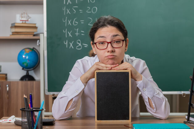 课桌年轻的女老师戴着眼镜拿着小黑板讲课 看起来很累 坐在教室黑板前的课桌旁上课老师坐着年轻