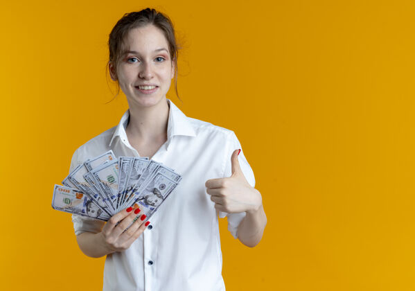 女孩年轻微笑的金发俄罗斯女孩拿着钱竖起大拇指在橙色与复制空间金发空间俄语