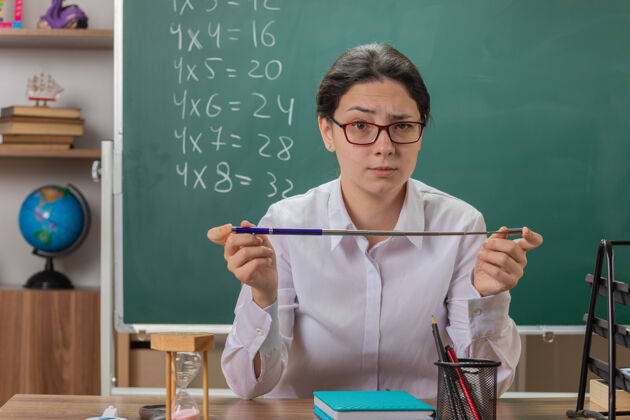 讲解年轻的女老师戴着眼镜 严肃地看着前面 拿着指针 坐在教室黑板前的课桌上讲解课文拿着戴着脸
