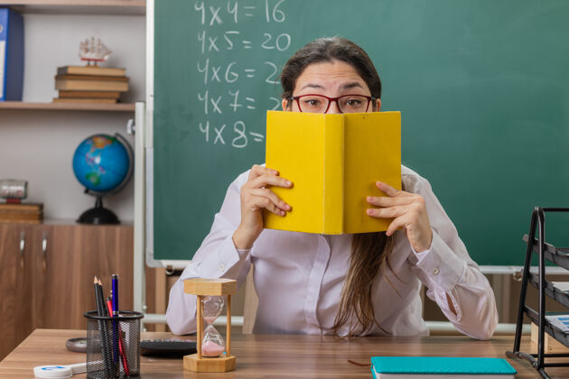 盖年轻的女老师戴着眼镜拿着一本书 用它遮住脸 坐在教室黑板前的课桌上备课教室女人眼镜