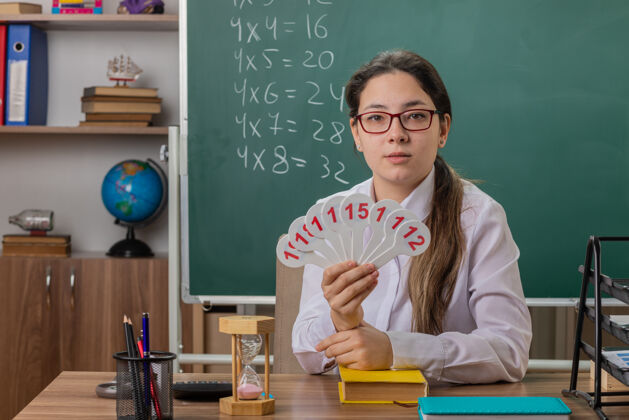 准备年轻的女老师戴着眼镜 手里拿着车牌 自信地坐在教室黑板前的课桌旁备课教室女人穿