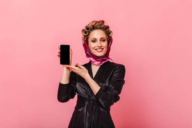 快乐一个穿着黑色长袍和围巾的积极的女人在粉红色的墙上展示智能手机衣服明亮美丽