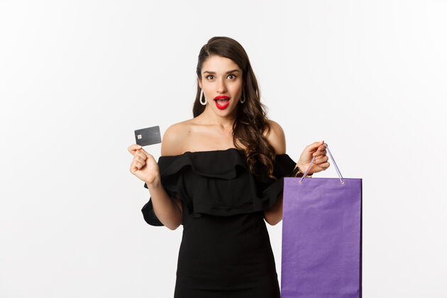 模型时尚的年轻女子穿着黑色衣服去购物 拿着包和信用卡 高兴地微笑着 站在白色的背景下黑发购物袋美丽