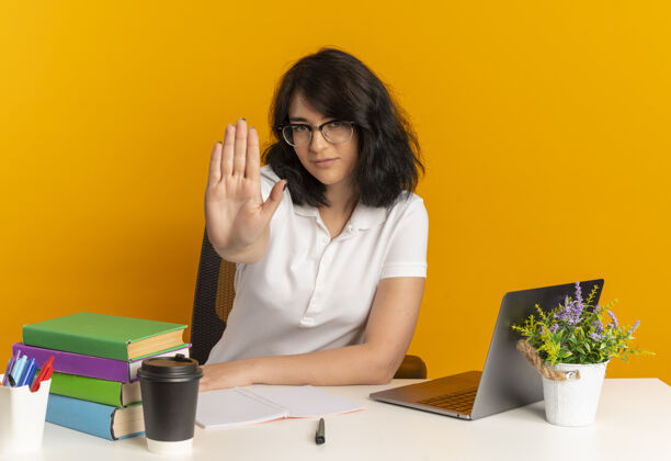 手年轻自信漂亮的白人女学生戴着眼镜坐在书桌旁 手里拿着学习工具 手势 手势 橙色 还有复印空间标志手势眼镜