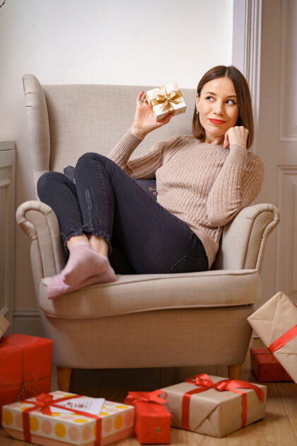 放松漂亮的年轻女子坐在舒适的扶手椅上 手里拿着一个礼品盒 周围都是礼物舒适家具年轻