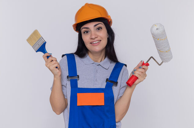 年轻快乐的年轻建设者妇女穿着建筑制服 戴着安全帽 拿着刷子和油漆辊 自信地站在白色的墙壁上 看着前面的samiling安全女人头盔