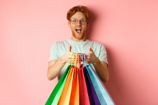 年轻快乐的红发男人买礼物 拿着购物袋 微笑着 站在粉红色的背景上红发礼物自信