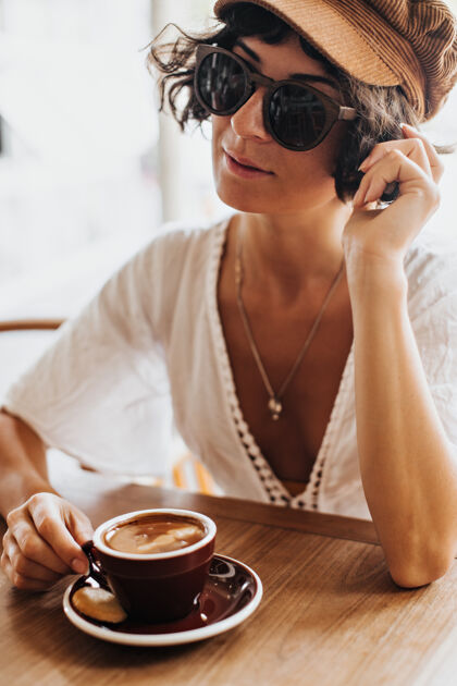 时尚棕褐色头发 戴着棕色帽子和太阳镜的女人拿着一个装着咖啡的棕色瓷杯 在咖啡馆里休息肖像异国情调黑发