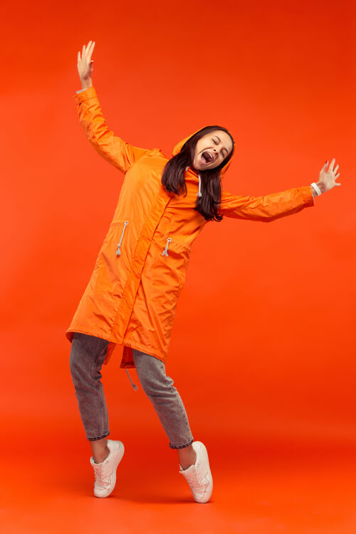 成人那个快乐微笑的小女孩穿着秋天的橘色夹克在摄影棚摆姿势 孤立在橘色的墙上快乐举行女性