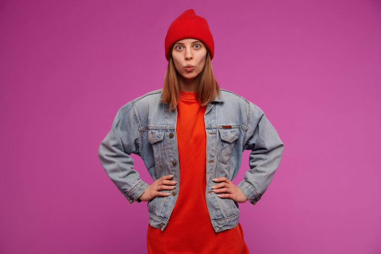 肖像少女 快乐的女人 深色长发穿着牛仔裤夹克 红色毛衣和帽子把手放在腰上 在紫色的墙上做亲吻的脸人面部学生