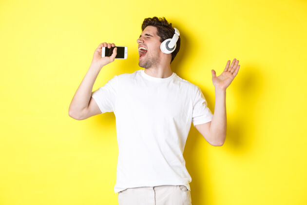 帅气快乐的家伙戴着耳机玩卡拉OK应用程序 对着智能手机麦克风唱歌 站在黄色背景上黄色成人听音乐