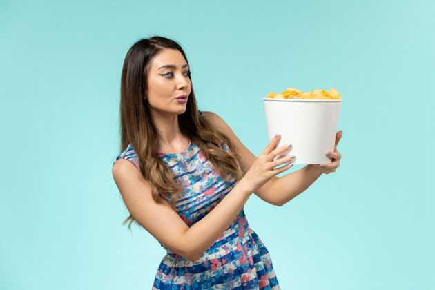 年轻的女性正面图：年轻的女性拿着篮子和薯片 在浅蓝色的表面上看电影电影院浅蓝色漂亮