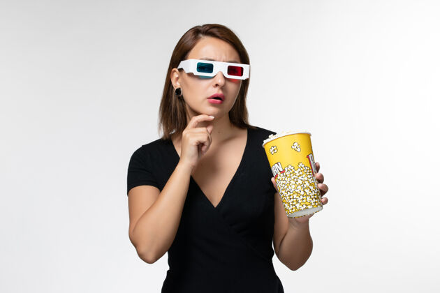 年轻女性正面图戴着d型太阳镜拿着爆米花的年轻女性在白色的表面上看电影和思考漂亮爆米花抱
