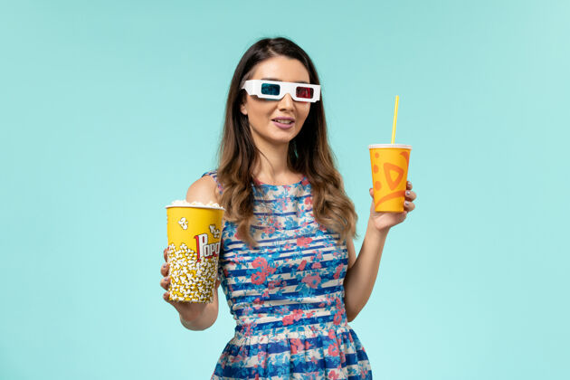 前面正面图蓝色表面上戴着d墨镜拿着爆米花饮料的年轻女性电影院漂亮电影院