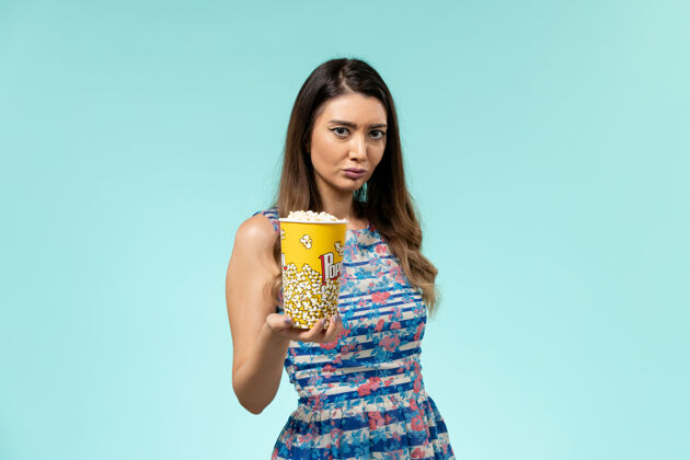 持有正面图年轻的女性拿着爆米花包在蓝色的表面上爆米花漂亮电影