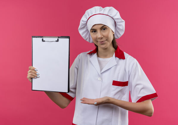 持有穿着厨师制服的年轻自信的白种人厨师女孩拿着并指着粉色空白处的剪贴板粉红厨师复制