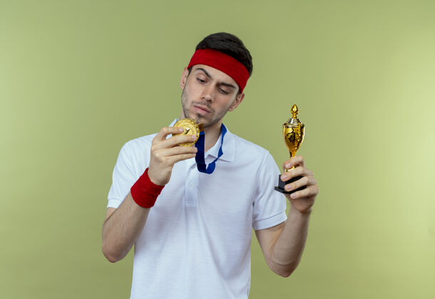 年轻戴着头巾 脖子上戴着金牌 站在绿墙上拿着奖杯的严肃的运动型年轻人严重男子头带