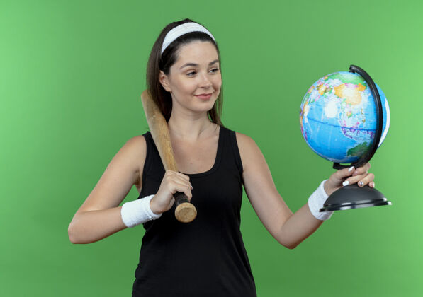 头带戴着头巾的年轻健身女士手持棒球棒和地球仪站在绿色的墙上 面带微笑地看着它微笑健身棒球