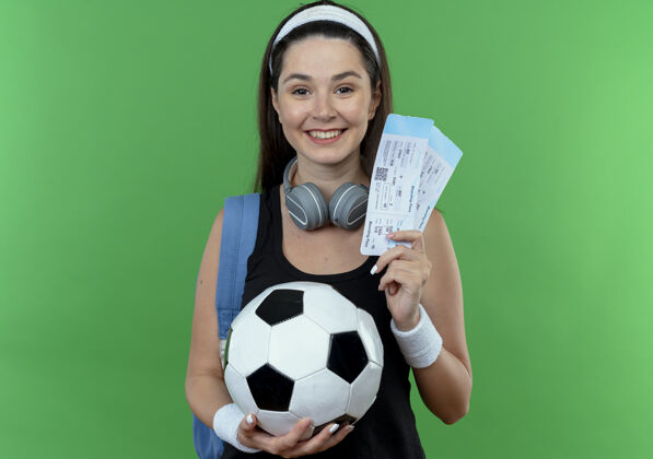微笑戴着头巾 背着耳机和背包 拿着足球和机票的年轻健身女士站在绿色的墙上 脸上洋溢着幸福的笑容举行立场年轻