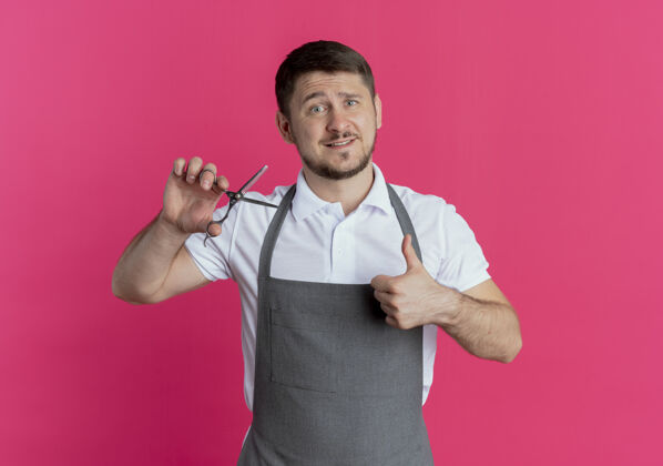 剪刀围裙上的理发师站在粉红色的墙上 手里拿着剪刀 竖起大拇指表演男人站着