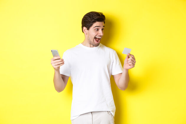 黄色惊讶的家伙拿着智能手机和信用卡 在黑色星期五网上购物 站在黄色背景下模特男人年轻
