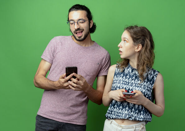 情侣一对年轻夫妇拿着智能手机 一个女人惊讶而困惑地看着自己的男朋友翻过绿墙男朋友惊喜年轻